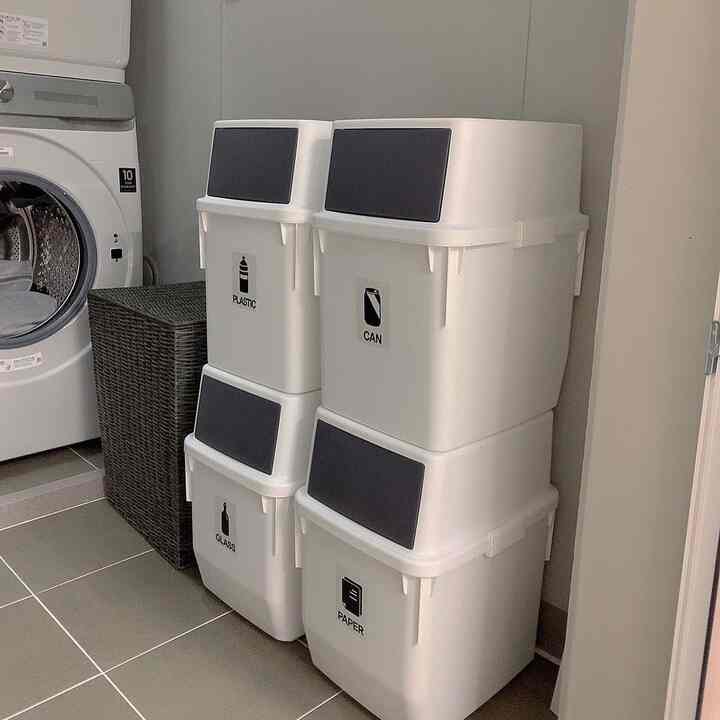 Clean laundry room featuring white tiled floor and washing machine with four stacked white recycling bins against beige wall