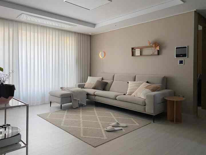 Natural beige-toned living room featuring beige sofa and rug, white chiffon curtains, and a wooden wall shelf creating a cozy atmosphere