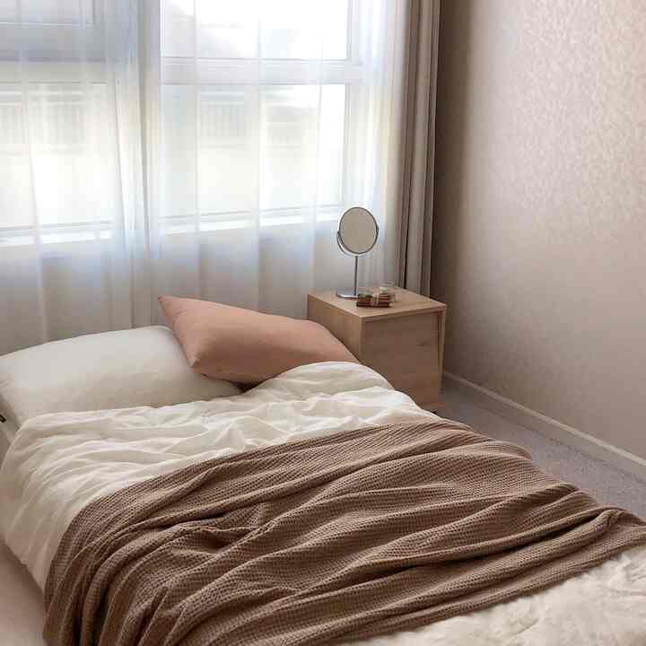 A white and beige toned bedroom featuring a simple bed, nightstand, and soft curtains creating a cozy atmosphere