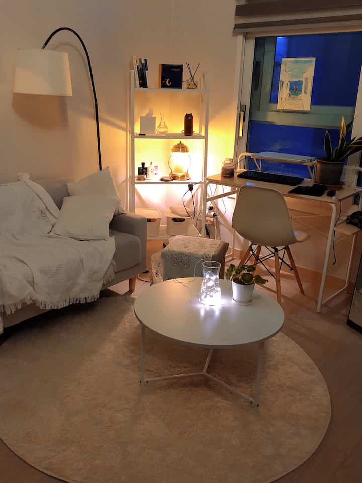 Cozy compact living room with warm lighting and white tones, featuring sofa and desk by window
