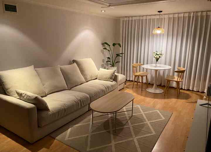 ベージュと白の色調のリビング、クリーム色のソファと木製のコーヒーテーブル、丸いダイニングテーブルとラタンのペンダントライトが配置された居心地の良い空間