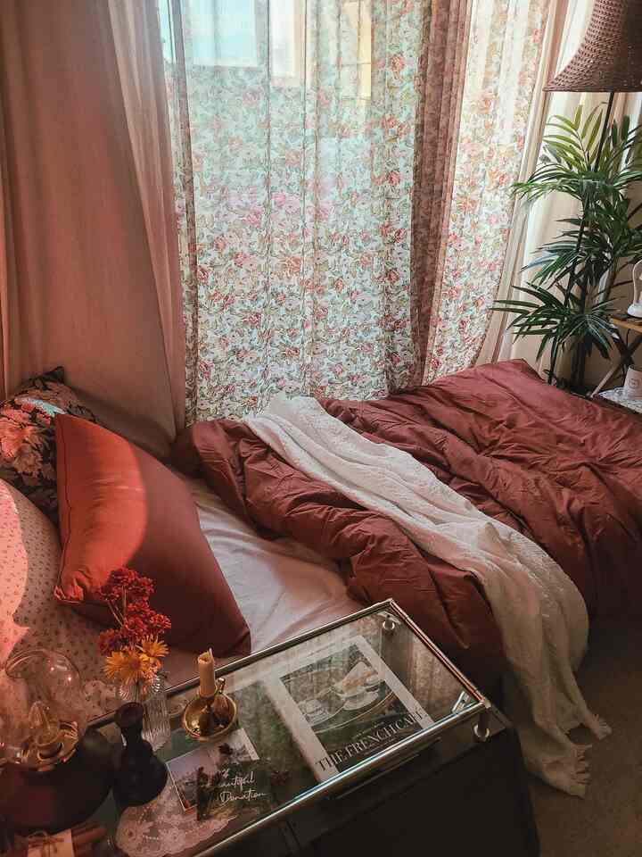 柔らかなブリックカラーと花柄カーテンが調和した寝室、植物とヴィンテージ感のあるインテリアが配置された雰囲気のある空間