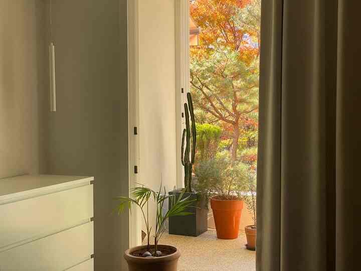 ナチュラルカラーの寝室ベランダ、チェストと遮光カーテン、多様な鉢植えが配置された落ち着いた空間