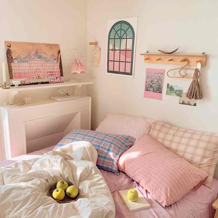 A cozy bedroom in pink and white tones featuring a comfortable bed with pillows, a decorative fireplace, and posters creating a natural atmosphere