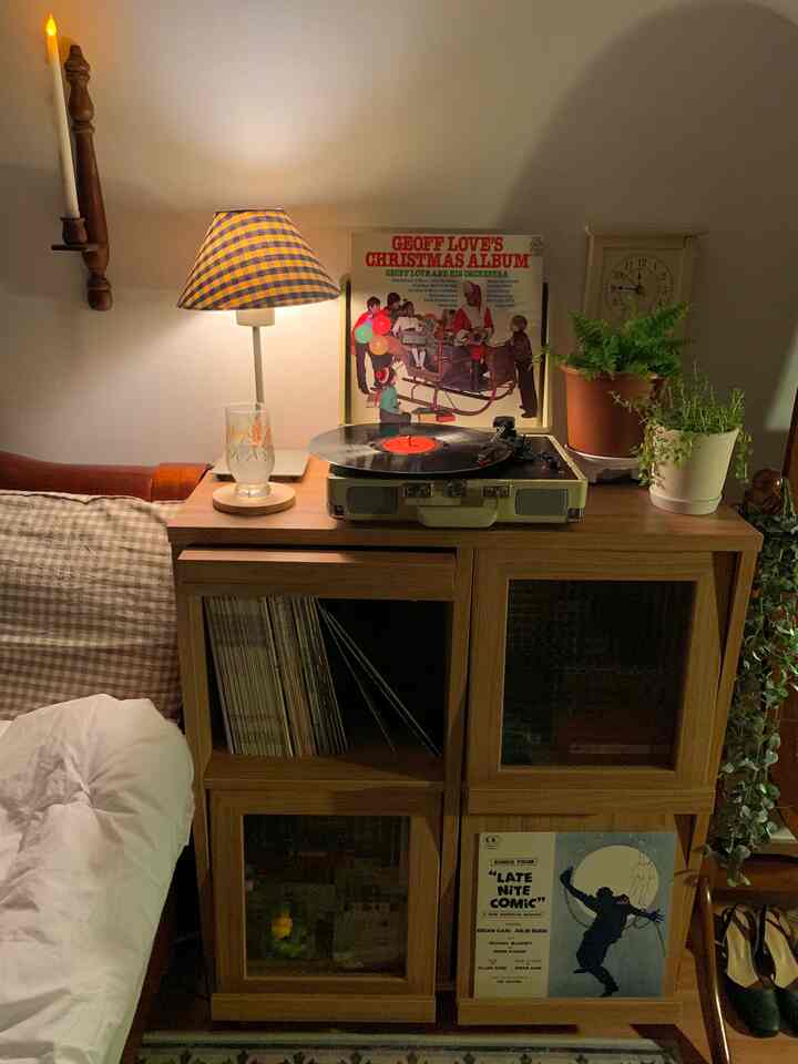 Cozy bedroom corner with warm lighting and natural wood-tone cabinet featuring vintage turntable, Christmas album, and plants