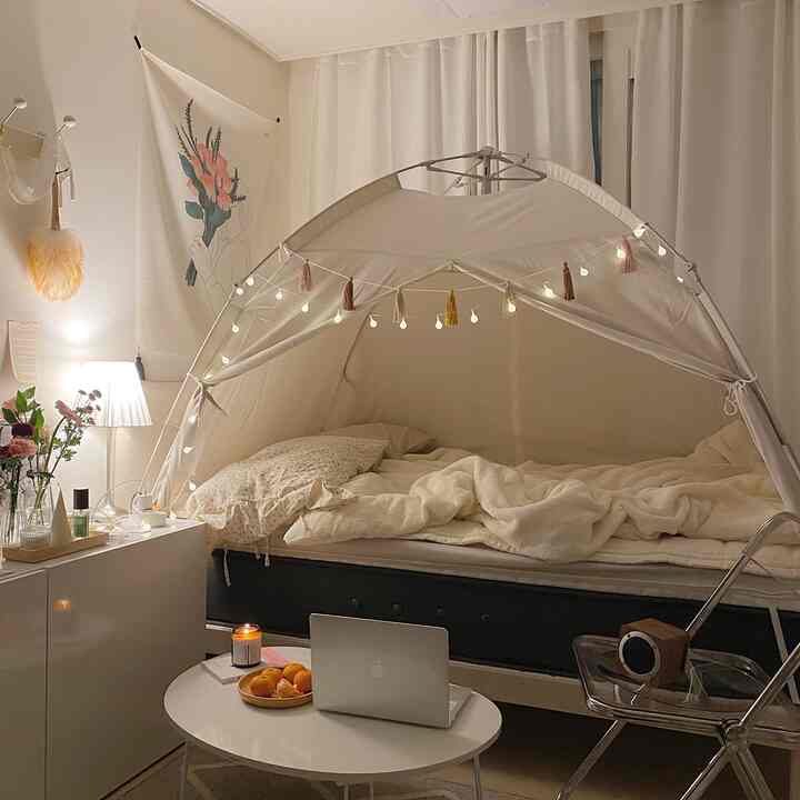 Cozy small bedroom in warm beige and white tones featuring a tent-style bed and a coffee table with a laptop