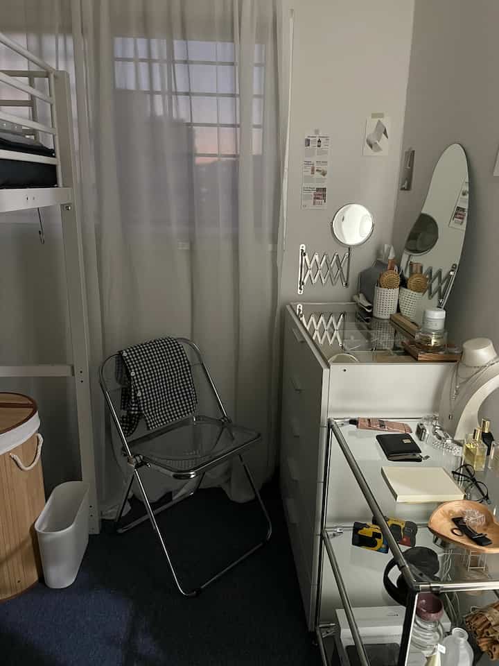 白を基調としたワンルームの寝室、透明な折りたたみ椅子と化粧台、棚が配置された実用的で居心地の良い個人空間