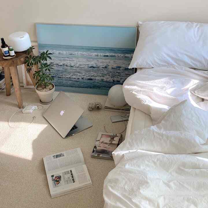 A natural-toned bedroom featuring white bedding, beige rug, ocean wave poster, and a potted plant creating a cozy atmosphere