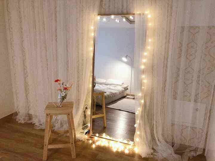 ウッドトーンの床と白いカーテンの寝室空間、大型鏡にLEDストリングライトが飾られた雰囲気のあるインテリア