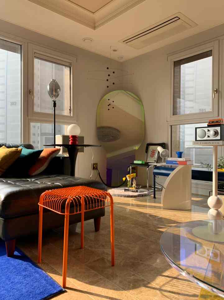 Beige-toned living room with natural sunlight, featuring black leather sofa, orange stool, and holographic mirror in a modern cozy setting