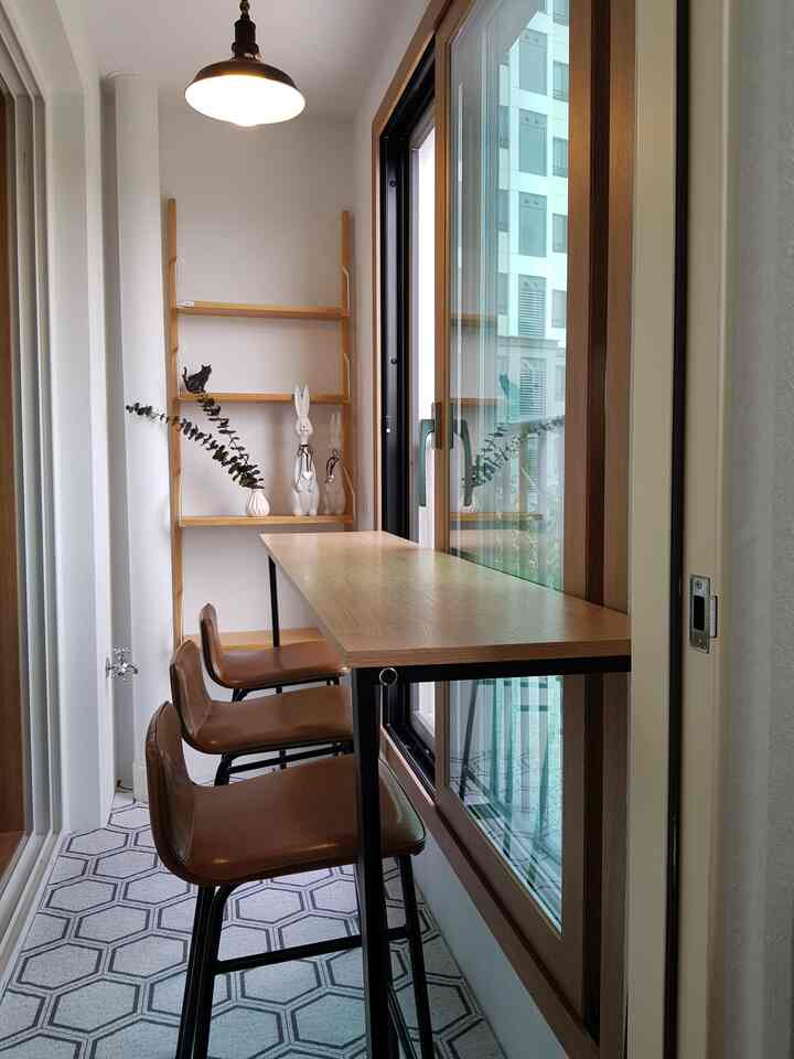 Natural wood tone bar table with three leather and steel bar stools in a narrow veranda with white walls, featuring a cozy home cafe atmosphere
