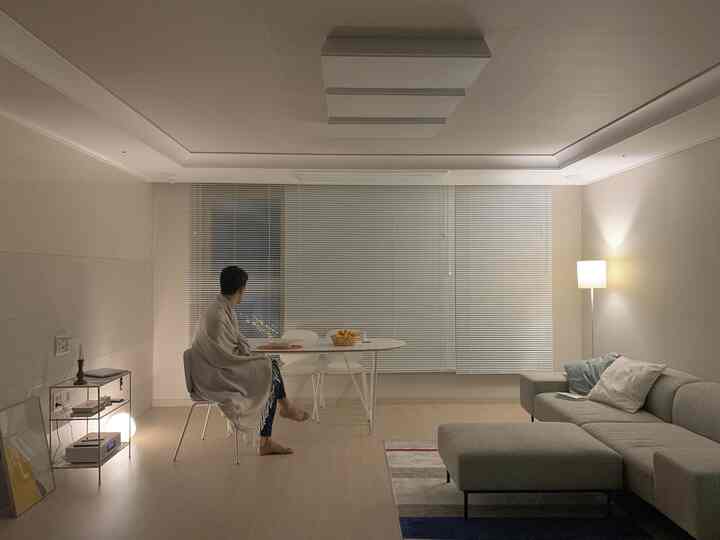 Natural color and white toned living room with gray sofa and white dining table, a man sits wrapped in blanket on dining chair looking out window