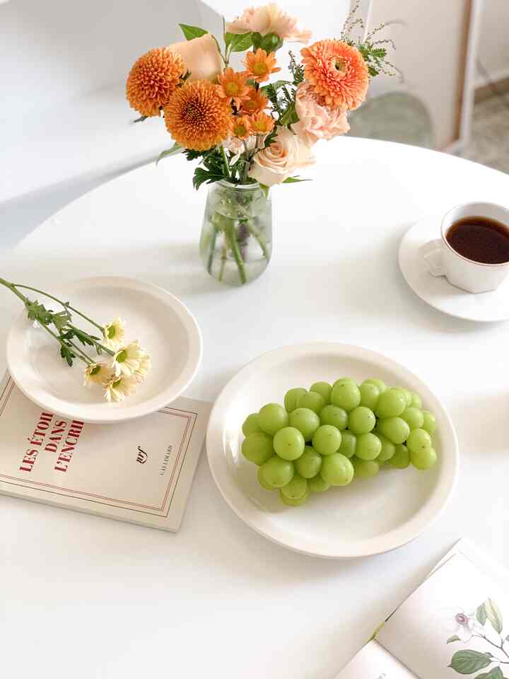白を基調としたキッチンテーブル、オレンジの花瓶とコーヒーカップ、ぶどうと皿が配置されたミニマルで自然な雰囲気のインテリア