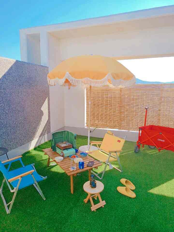 Bright yellow umbrella and chairs, wood-tone outdoor table with green grass carpet create a cozy rooftop terrace camping space