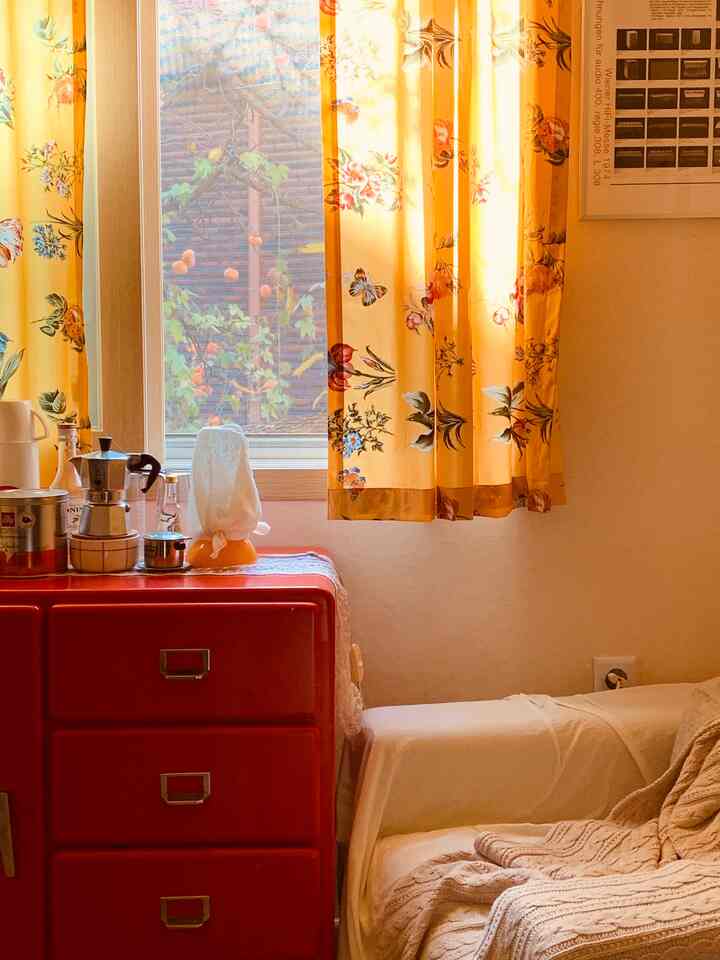 レッドのチェストの横にベージュのソファと黄色の花柄カーテンがある温かみのあるヴィンテージ風キッチン空間