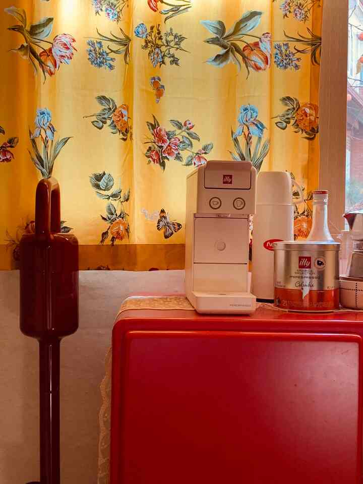 Warm-toned home cafe space featuring yellow floral curtains and a red cabinet with a white coffee machine and coffee capsules on top
