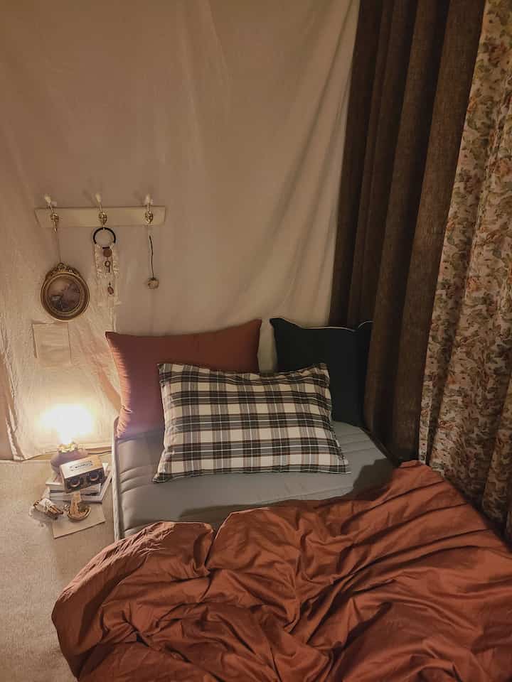 茶色とグレーのトーンが調和する小さな寝室、メモリーフォームマットレスと複数の枕、花柄カーテンが配置された雰囲気のあるヴィンテージ空間