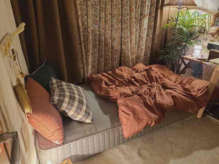 暖かいベージュとブラウンのトーンが特徴の小さな寝室、ふかふかのマットレスとフロアランプ、植物が配置された雰囲気のあるインテリア