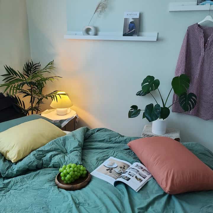 A soft green and natural-toned bedroom featuring green plants, bedding, and warm lighting creating a cozy atmosphere