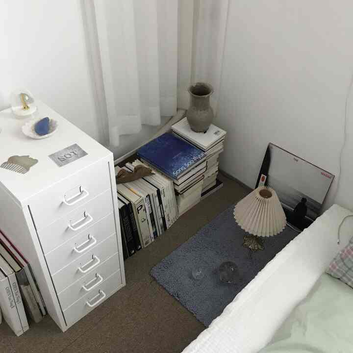 A minimal natural bedroom corner in white and blue tones featuring a dresser, stacks of books, a rug, and a table lamp