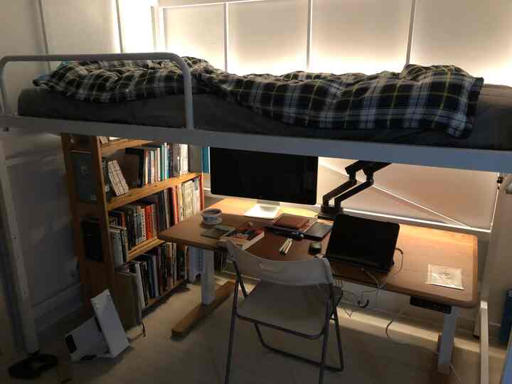 A compact single-person room in white and wood tones featuring a bunk bed with desk and bookshelf underneath, creating a practical workspace