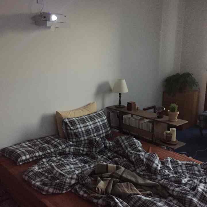 Natural modern bedroom with white walls and brown bed frame, featuring wall-mounted projector, bookshelf, and table lamp