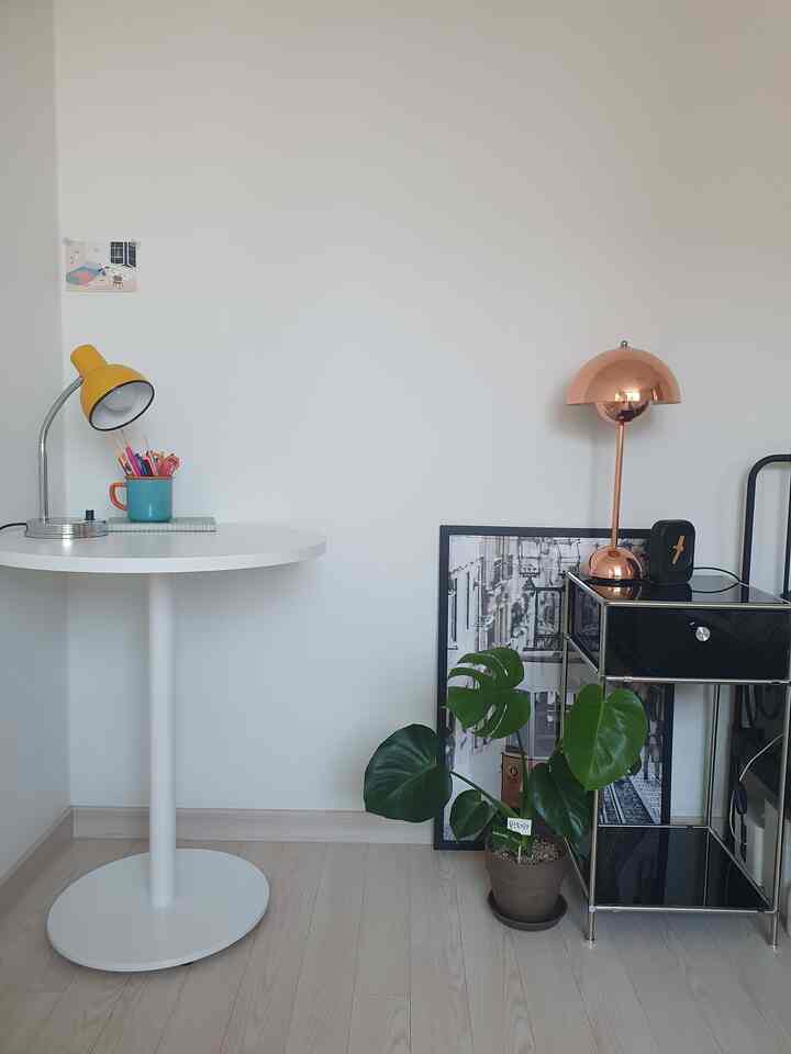 ホワイトの壁と円形テーブル、グリーンのモンステラの鉢植え、銅色のテーブルランプがあるすっきりとしたコーナー収納の空間