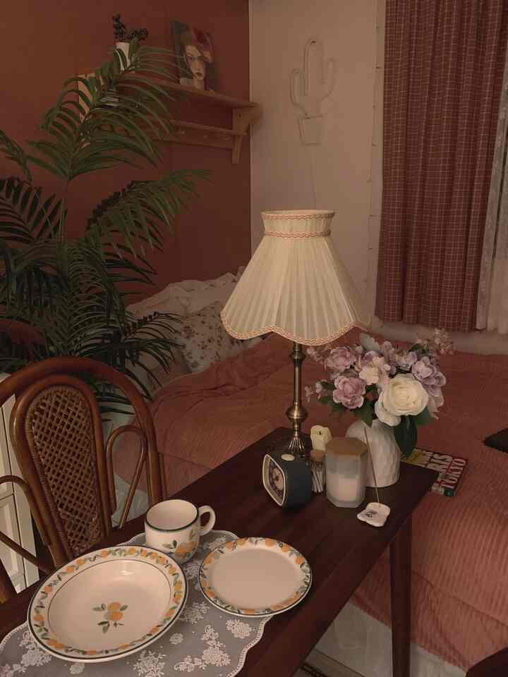 ピンクトーンの寝具とカーテンが印象的なヴィンテージスタイルの寝室で、木製家具と大きな植物が配置された居心地の良い空間