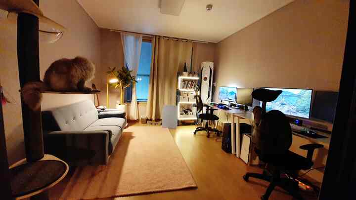 黄色照明とベージュカーテンのあるワンルーム作業スペース、デュアルモニターの机、ソファ、棚が配置された落ち着いた空間