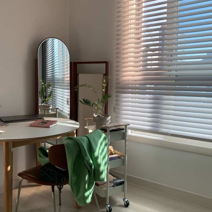 A cozy small living room with natural wood and white tones featuring a round table, full-length mirror, and blinds