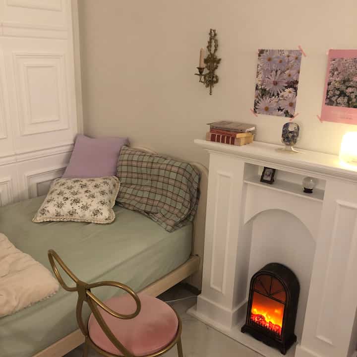 A cozy bedroom featuring a mint-colored bed with pink cushions and a white decorative fireplace