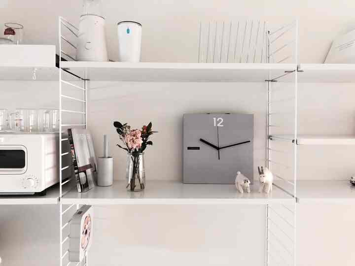 ホワイトとグレーのトーンのキッチンスペース、ストリングシェルフと壁時計、花瓶が配置されたミニマルなインテリア