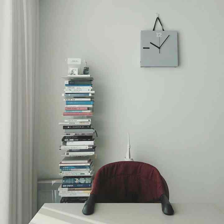 Modern space with white walls and table featuring a tall bookshelf stacked with books on the left and a wall clock on upper right