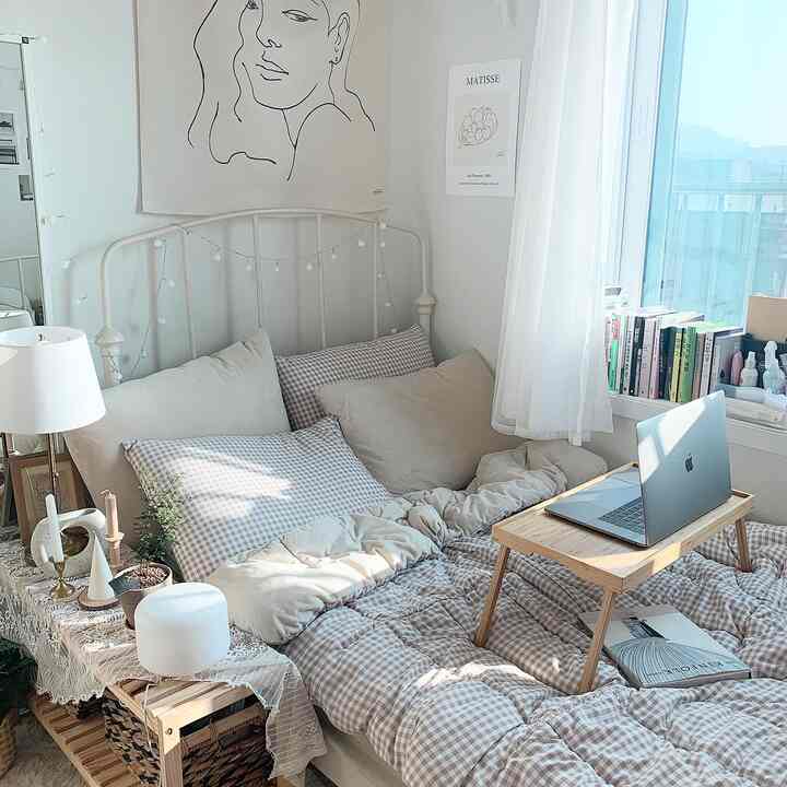 Natural-toned studio bedroom featuring a metal bed frame, wooden laptop table, and blackout curtains creating a cozy atmosphere