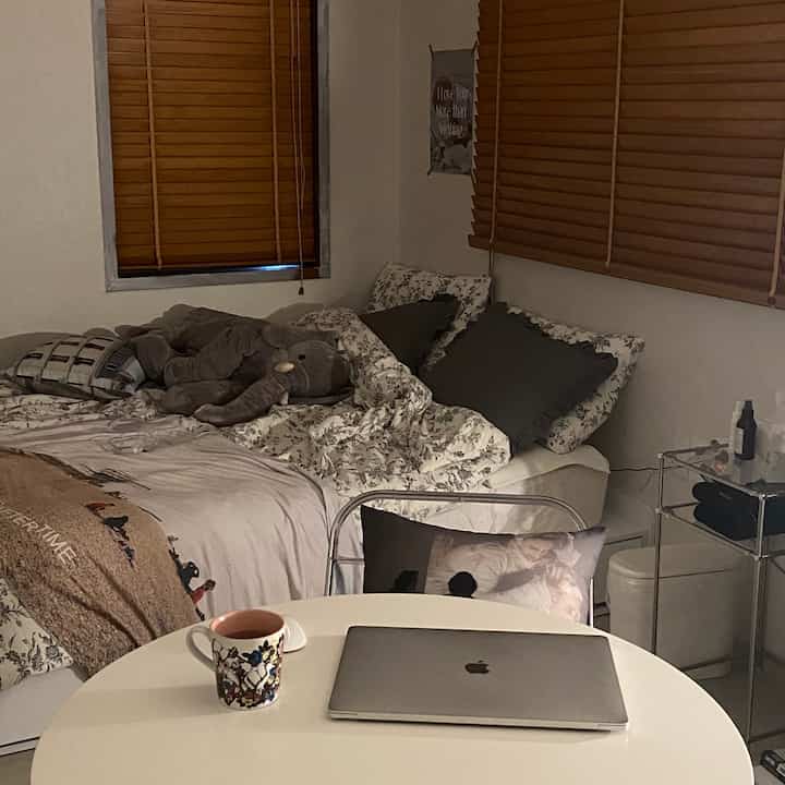 White and wood-tone curtained bedroom featuring a bed and a round table with laptop and mug creating a cozy atmosphere