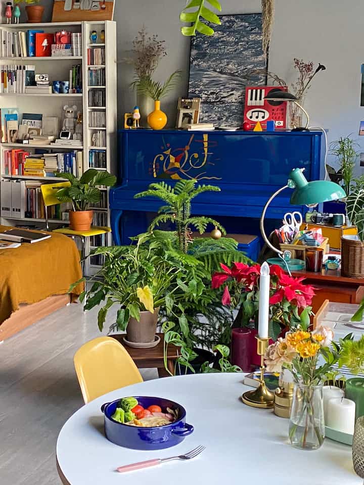 Cozy studio dining space with a blue piano, yellow chair, and various plants arranged around a white round table