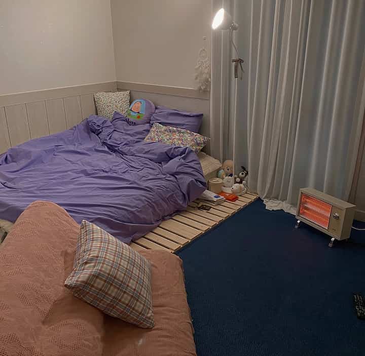 紫の寝具が印象的な小さなワンルーム寝室、ネイビーカーペットと暖かい電気ヒーターが配置された居心地の良い空間