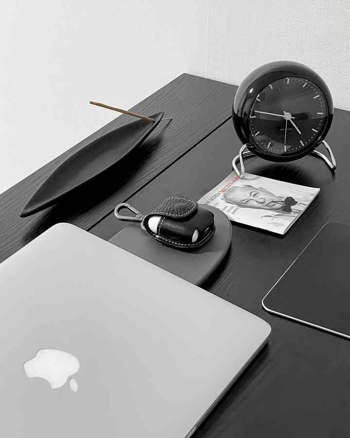 黒い木製デスクの上にミニマルでモノトーンなスタイルのワイヤレス充電器、エアポッズケース、卓上時計、香立てが整然と配置された空間