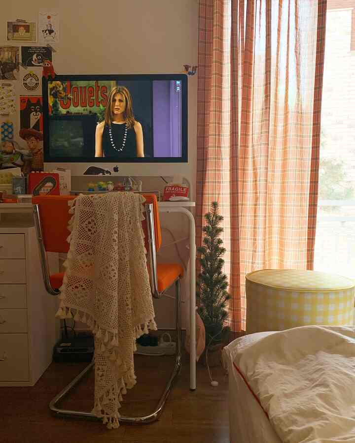 Orange-toned bedroom featuring a desk with monitor, checkered curtains, and a yellow stool creating a cozy atmosphere