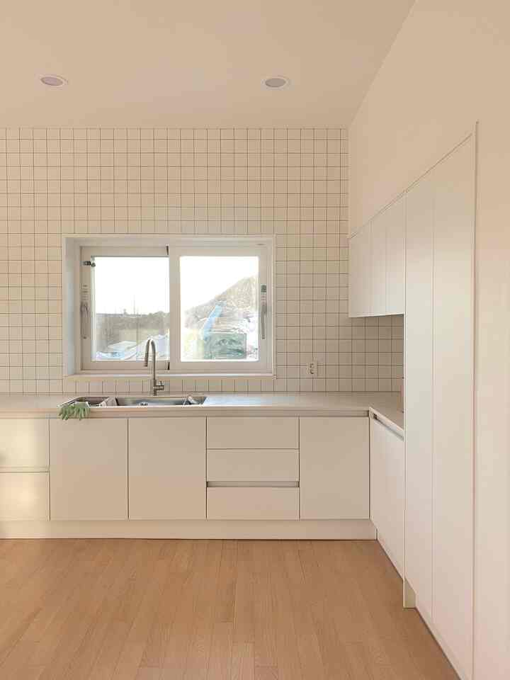 A simple white-toned kitchen with wooden flooring, featuring cabinets placed on one side for an open and airy feel