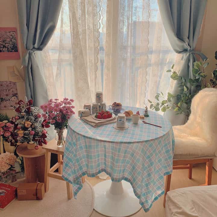 Cozy home cafe space with soft blue and white tones featuring a round table, lace curtains, and wood furniture in a compact room