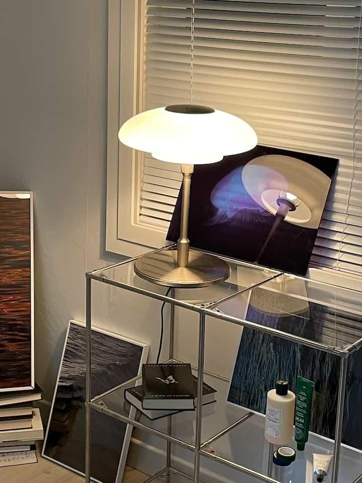 Modern bedroom corner featuring white blinds, steel-based modular shelving with a table lamp and several posters