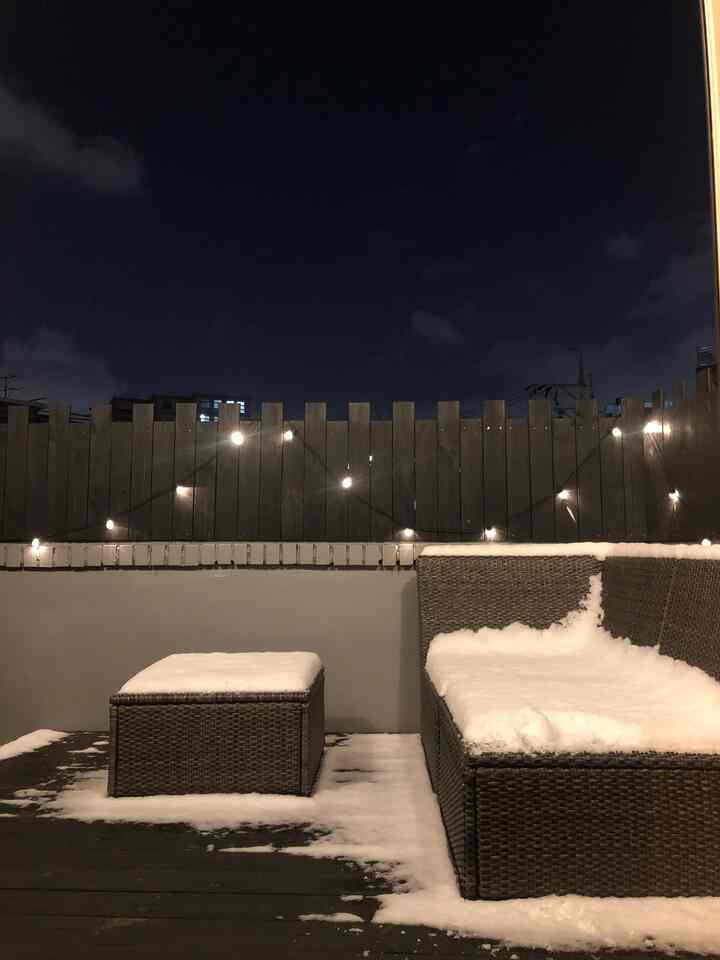 Dark night rooftop terrace in brown wood tones covered with snow, featuring string lights and cozy outdoor sofa seating