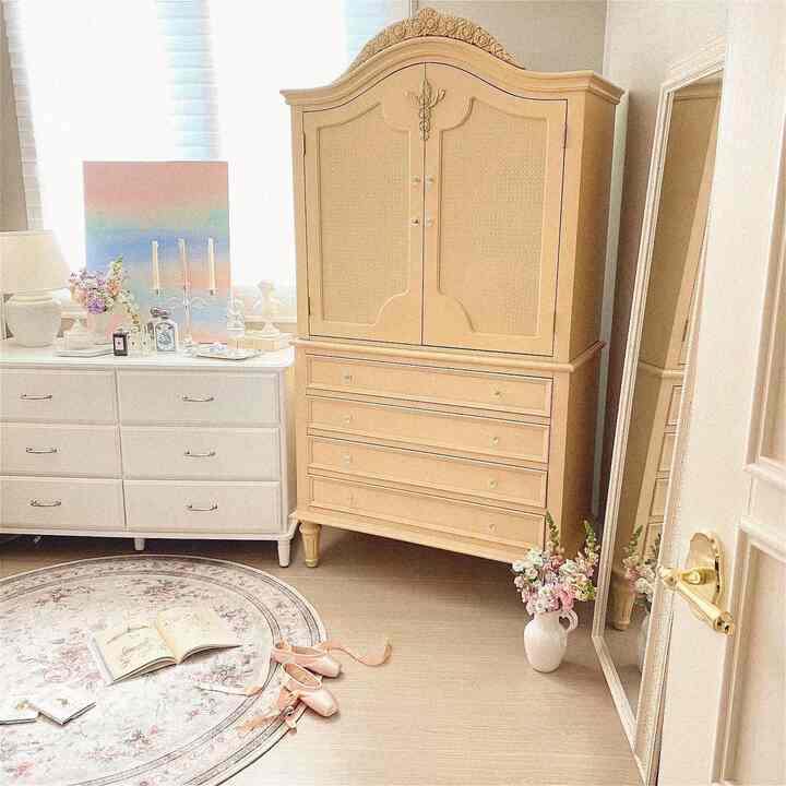 Pastel-toned walk-in closet space featuring a beige wooden wardrobe, white dresser, flowers and a decorative rug creating a cozy atmosphere