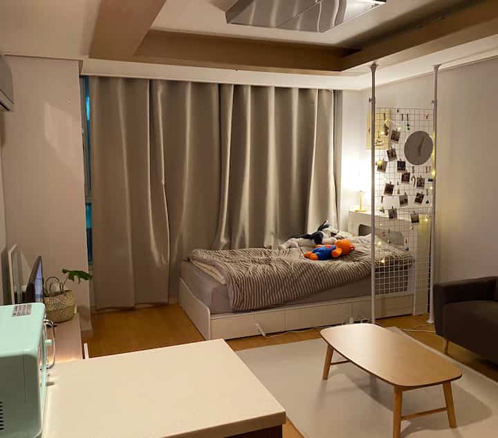 Beige and white toned studio apartment featuring bed, curtains, and sofa with a cozy, natural atmosphere