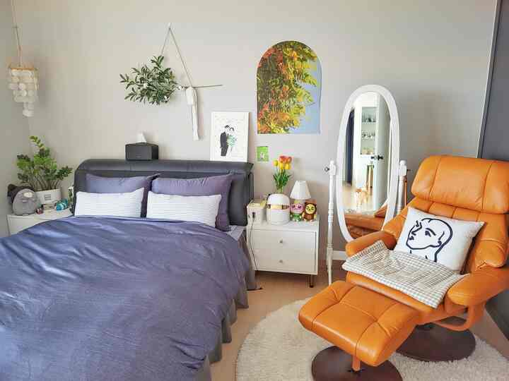 Gray-walled bedroom featuring an orange recliner, arched poster, full-length mirror, nightstand, and bedding creating a cozy atmosphere