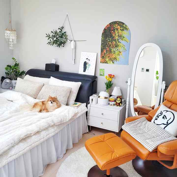 White bedroom with a large bed center-left featuring a resting dog, and a bright orange armchair by a standing mirror, cozy natural modern vibe