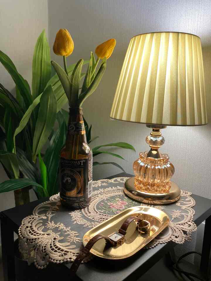 グレーの壁と黒いナイトテーブルの上に、ゴールドのテーブルランプとトレイ、黄色のチューリップの造花が置かれた雰囲気のある寝室のナイトテーブル