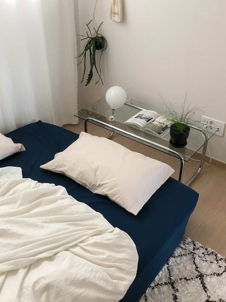 ネイビーとホワイトのトーンの寝室、ネイビーマットレスカバーと白い寝具、サイドにガラスのサイドテーブルと小さな植物が置かれたミニマルでナチュラルなインテリア