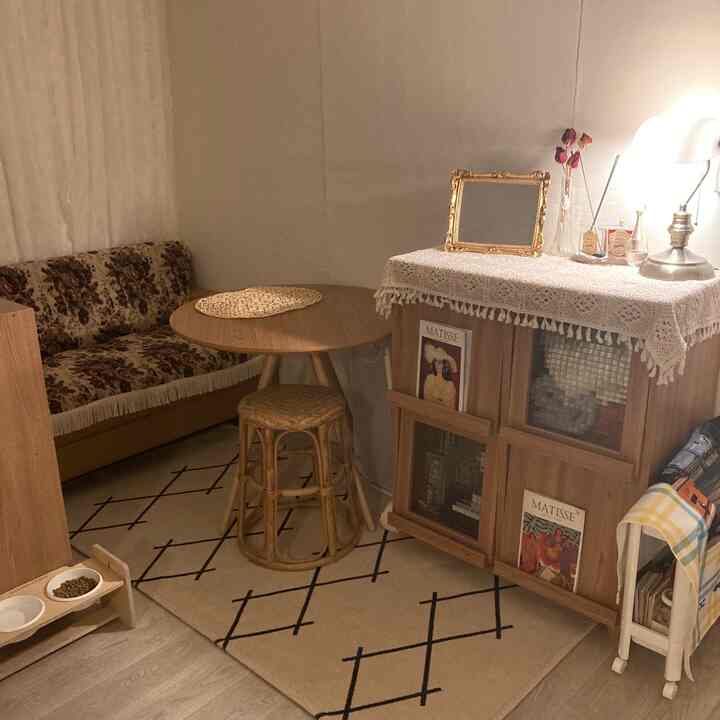 ベージュとブラウンのトーンのワンルームリビング、ヴィンテージ柄のソファ、円形テーブル、ラタンのスツール、レースカーテンと化粧台が配置された暖かみのある雰囲気の空間